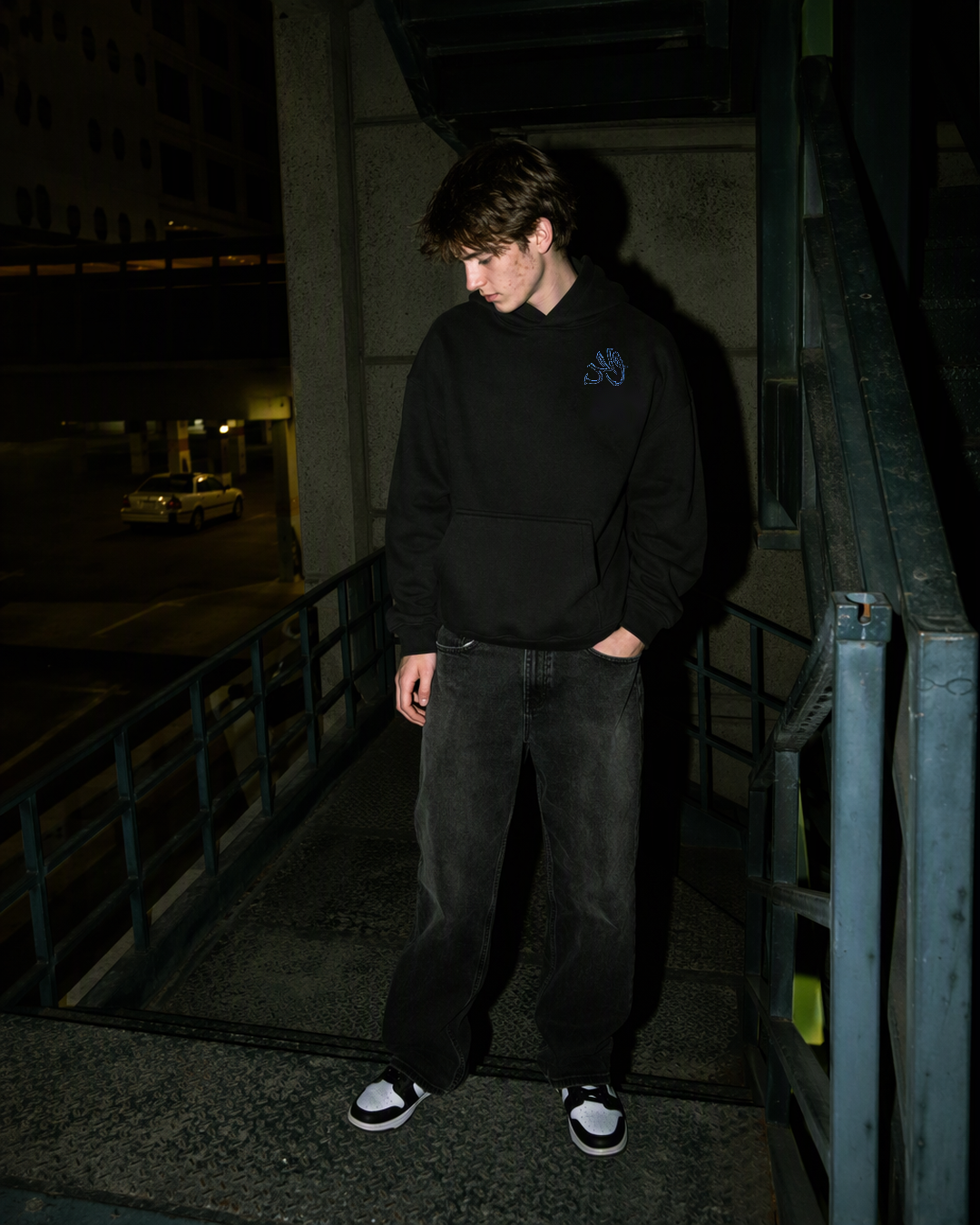 Digitally created campaign portrait of an adult model wearing the Black Forest Wahid hoodie beside an outdoor stair railing.